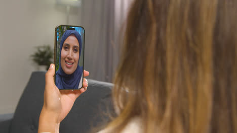 Close-Up-Of-Woman-Sitting-On-Sofa-At-Home-Having-Video-Call-With-Female-Muslim-Friend-On-Mobile-Phone